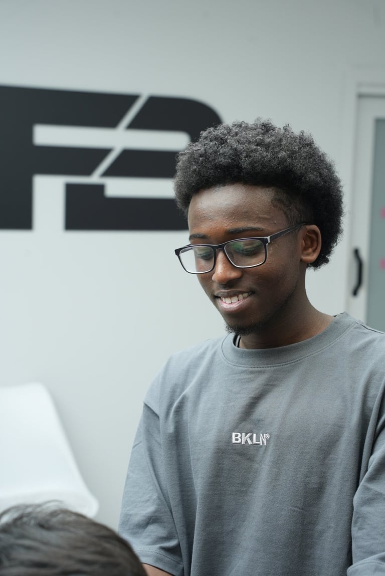 Young man wearing glasses and gray BKLYN shirt smiling indoors with modern geometric sign in background